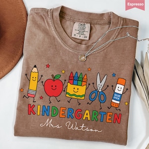May include: An espresso-colored t-shirt featuring a graphic of a smiling pencil, apple, crayon box, scissors, and glue stick. The text reads KINDERGARTEN and Mrs. Watson. The shirt has a Comfort Colors label and a silver-toned necklace rests on it.