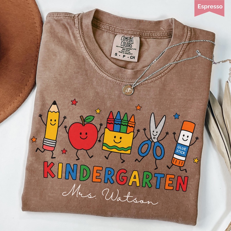 May include: An espresso-colored t-shirt featuring a graphic of a smiling pencil, apple, crayon box, scissors, and glue stick. The text reads KINDERGARTEN and Mrs. Watson. The shirt has a Comfort Colors label and a silver-toned necklace rests on it.