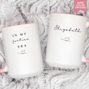 May include: Two white ceramic mugs with pink handles and rims. One mug reads "IN MY forties ERA" with a heart, the other has the name "Elizabeth" and a heart. Each mug is 15 oz.