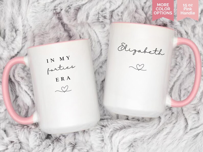 May include: Two white ceramic mugs with pink handles and rims. One mug reads "IN MY forties ERA" with a heart, the other has the name "Elizabeth" and a heart. Each mug is 15 oz.