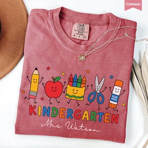 May include: A crimson short-sleeved t-shirt featuring a graphic of a smiling pencil, apple, crayon box, scissors, and glue stick. The text reads KINDERGARTEN and Mrs. Watson. The shirt has a Comfort Colors label at the neck and a small silver necklace.