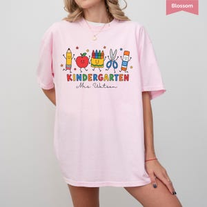 May include: A light pink t-shirt featuring a colorful graphic of a pencil, apple, crayon box, scissors, and glue stick with faces. The text reads KINDERGARTEN and Mrs. Watson in a script font. Small stars are scattered around the school supplies.