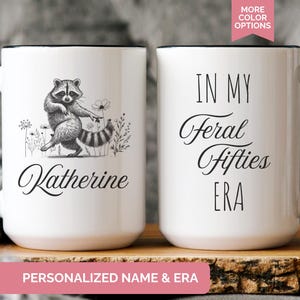 White ceramic mug featuring a hand-drawn raccoon holding a flower on one side with personalized name, and the phrase “In My Feral Fifties Era” on the other.