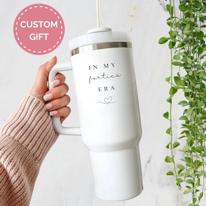 May include: White tumbler with a handle, featuring the text "IN MY forties era" and a heart symbol. A pink circle with the words "CUSTOM GIFT" is in the upper left. The tumbler is held by a person wearing a light pink sweater.