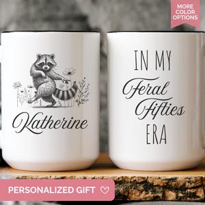 White ceramic mug featuring a hand-drawn raccoon holding a flower on one side with personalized name, and the phrase “In My Feral Fifties Era” on the other.