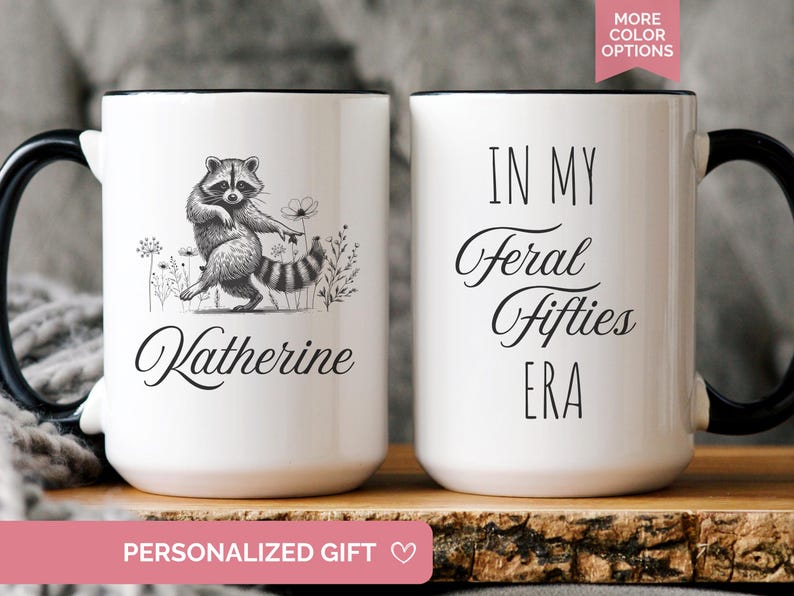 White ceramic mug featuring a hand-drawn raccoon holding a flower on one side with personalized name, and the phrase “In My Feral Fifties Era” on the other.