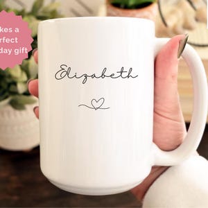 May include: A white 15 oz ceramic mug with the name "Elizabeth" in a cursive font and a heart symbol. The mug has a large handle and is held by a person. Text on the mug reads "Makes a perfect birthday gift".