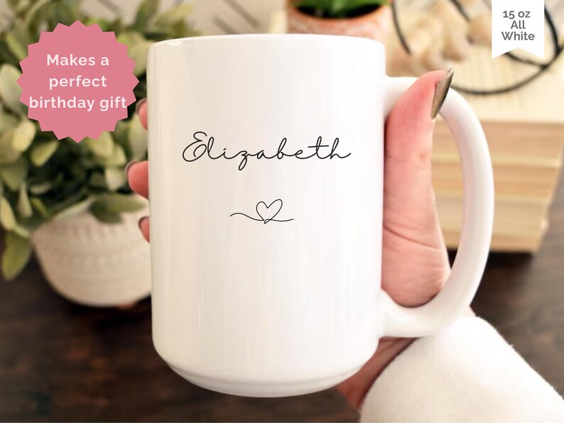 May include: A white 15 oz ceramic mug with the name "Elizabeth" in a cursive font and a heart symbol. The mug has a large handle and is held by a person. Text on the mug reads "Makes a perfect birthday gift".