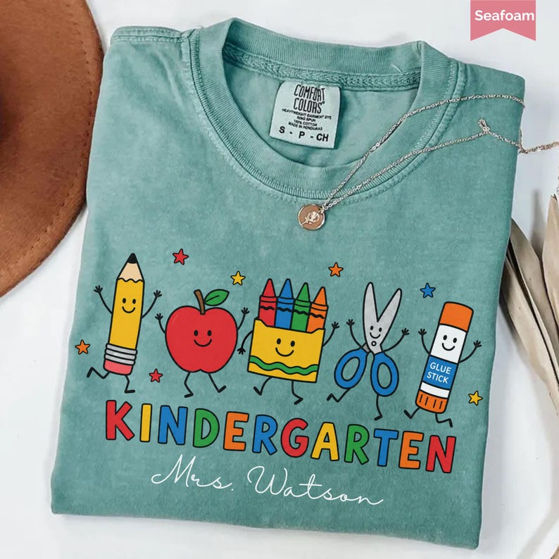 May include: A seafoam green t-shirt featuring a graphic of a smiling pencil, apple, crayon box, scissors, and glue stick. The text reads KINDERGARTEN and Mrs. Watson. The shirt has a Comfort Colors label and is paired with a delicate gold necklace.