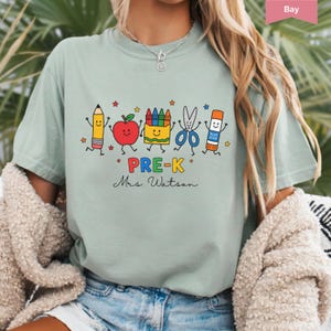 May include: A sage green t-shirt featuring a graphic of a pencil, apple, crayon box, scissors, and glue stick with smiling faces. The text reads PRE-K and Mrs. Watson in black script. Small colorful stars are scattered around the school supplies.