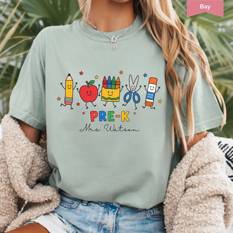 May include: A sage green t-shirt featuring a graphic of a pencil, apple, crayon box, scissors, and glue stick with smiling faces. The text reads PRE-K and Mrs. Watson in black script. Small colorful stars are scattered around the school supplies.