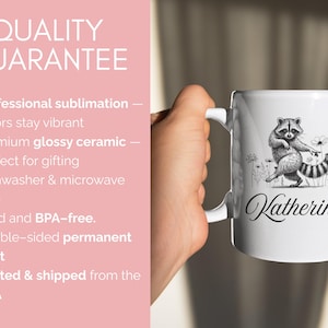 May include: White ceramic mug featuring a black and white illustration of a raccoon with a flower and the name "Katherine" in cursive. The mug has a white handle and is held by a person.
