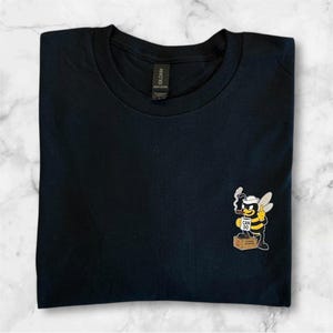 Può includere: T-shirt nero con una grafica a fumetto di un'ape. L'ape indossa un cappello e tiene in mano un sigaro, con le parole "CAN DO" sulla scatola su cui è in piedi. La maglietta è piegata ordinatamente.