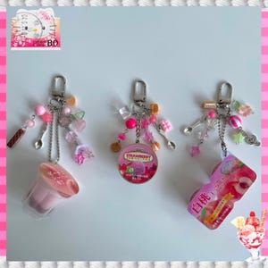 以下が含まれることがあります： 銀色の留め金が付いた3つのキーホルダー。それぞれピンクとクリアのビーズ、ミニチュアのスプーン、チャームで飾られています。1つのキーホルダーにはピンクのデザートカップ、もう1つにはイチゴのアイスクリーム容器、3つ目には桃のヨーグルト容器が付いています。