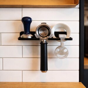 Könnte beinhalten: Eine schwarze Espresso-Station mit einem blauen Tamper, einem Siebträger und einem weißen Ring. Ein lächelnder, gesichtsähnlicher Aufsatz hängt darunter. Die Artikel sind an einer weißen gefliesten Wand montiert.