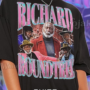May include: Black unisex t-shirt featuring a graphic of Richard Roundtree with the text "RICHARD ROUNDTREE" in pink and teal. The shirt has a vintage aesthetic and includes the word "Unisex".