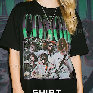 May include: Black unisex t-shirt featuring a band graphic with the name "Conor Oberst" in a green and purple design. The shirt includes a photo of the band members and the text "Shirt Unisex".