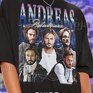 Op de afbeelding: Zwart unisex T-shirt met een afbeelding van Andreas Pietschmann en zijn naam in blauw en wit. Het shirt bevat ook de tekst "SHIRT Unisex" en maat informatie.