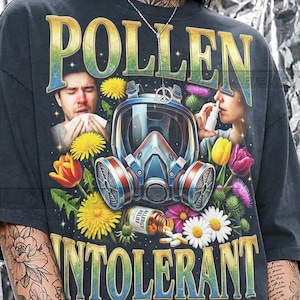 May include: Dark gray t-shirt with the words "POLLEN INTOLERANT" in a vintage style. The graphic includes a gas mask, flowers, and people experiencing allergy symptoms. The shirt also has the words "DENIM COMFORT COLORS".