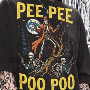 May include: Dark gray t-shirt with a graphic featuring a skeleton cowboy holding two guns, standing on a tree, with two skeletons below. The text "PEE PEE POO POO" is printed in yellow.