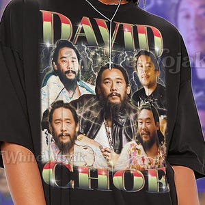 May include: Black unisex t-shirt featuring a graphic of a man's face with the text "DAVID CHOE". The shirt has a vintage style with lightning bolt accents. The text "SHIRT Unisex" is also printed on the shirt.
