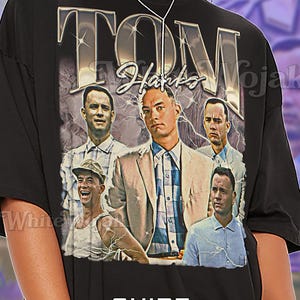 May include: Black unisex t-shirt featuring a graphic of Tom Hanks with the text "TOM Hanks". The design includes multiple images of the actor. The shirt also has the word "Unisex" printed on it.