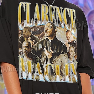 May include: Black unisex t-shirt featuring a graphic design with the text "CLARENCE MACLIN" in gold lettering. The design includes images of several men. The shirt also has the word "Unisex" printed on it.