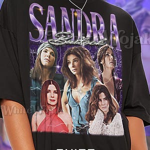 May include: Black unisex t-shirt featuring a graphic of Sandra Bullock with the text "SANDRA Bullock" in purple. The shirt has a vintage style with multiple images of the actress. The text "SHIRT Unisex" is also visible.