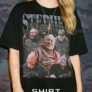 May include: Black unisex t-shirt featuring a graphic of multiple images of a man with the text "Stevie McKinley Henderson". The shirt also has the word "Shirt" and "Unisex" printed on it. The model is wearing a 2XL shirt.