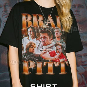 Può includere: T-shirt unisex nera con la scritta "BRAD PITT" in oro e un collage di immagini. La maglietta ha anche la parola "SHIRT" sopra "Unisex" in bianco. La modella indossa una maglietta 2XL.