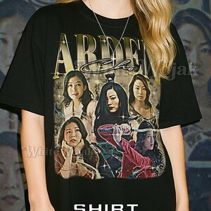 Puede incluir: Camiseta unisex negra con un gráfico de seis mujeres en un diseño estilizado. La camiseta tiene el texto "ARDEN Cho" y "SHIRT Unisex" impreso. La modelo lleva una camiseta 2XL.