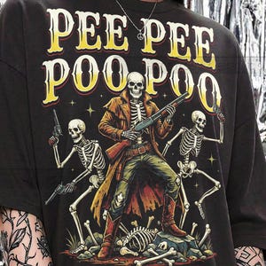 May include: Dark gray t-shirt with the text "PEE PEE POO POO" in yellow and red. The graphic features a skeleton cowboy holding a shotgun, flanked by two skeletons with pistols. The shirt is labeled "GRAPHITE COMFORT COLORS."