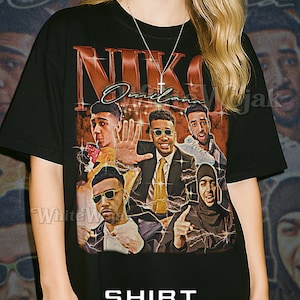 May include: Black unisex t-shirt featuring a graphic of a group of men with the text "Niko Omilana" in a vintage style. The shirt also has the word "Shirt Unisex" printed in white.