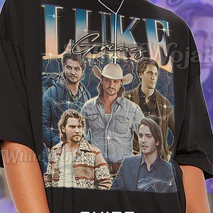 May include: Black unisex t-shirt featuring a graphic of Luke Grimes with text. The shirt has a vintage design with a lightning bolt effect. The text "SHIRT Unisex" is at the bottom.