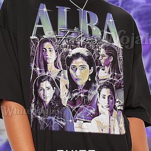 May include: Black unisex t-shirt featuring a graphic design with the word "ALBA" in a stylized font, along with images of women. The shirt has the word "Shirt" and "Unisex" printed on it.