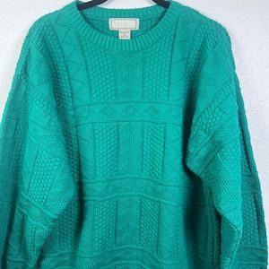 May include: A vibrant emerald green long-sleeve sweater featuring a complex knit pattern with vertical columns of cable and textured stitches. The garment has a crew neckline and a label at the inner collar that reads Christopher Hayes.