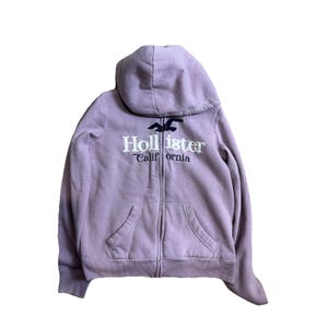 May include: A light purple zip-up hoodie featuring a hood, long sleeves, and two front pockets. The chest displays the dark blue embroidered text Hollister California with a dark blue bird logo positioned above the word Hollister in white.