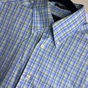 May include: A button-down shirt featuring a plaid pattern in shades of light blue, white, and lime green. The shirt has a pointed collar with buttons at the tips, a front button placket with white buttons, and a label inside the collar reading Orvis.