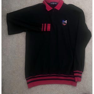 May include: Black long-sleeved shirt with a red collar and cuffs. The shirt features a quarter-zip closure and a crest on the chest. The cuffs and hem have red stripes. The brand name "Ralph Lauren" is visible on the collar.