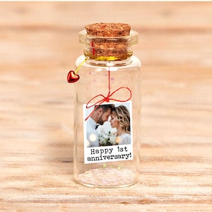 Può includere: Una piccola bottiglia di vetro con tappo di sughero, contenente glitter e una foto con la scritta "Happy 1st anniversary!". Un ciondolo a forma di cuore rosso è appeso al collo della bottiglia. La foto mostra una coppia.