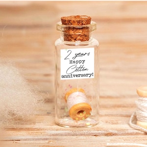 May include: A small glass bottle with a cork stopper contains a miniature spool of thread. The bottle has a label that reads "2 years Happy Cotton anniversary!". Two additional spools of thread sit nearby. The background is a light wood surface.