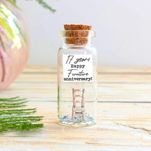 Op de afbeelding: Een doorzichtige glazen fles met een kurkstop die een miniatuur metalen stoel en zilveren glitters bevat. Een wit label op de voorkant van de fles bevat de zwarte tekst 17 years Happy Furniture anniversary! De fles staat rechtop.