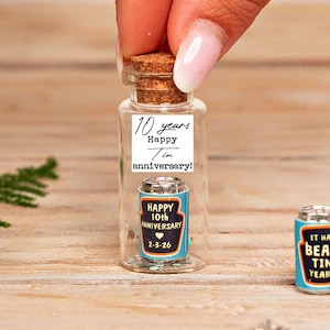 Pode incluir: Uma pequena garrafa de vidro transparente com rolha de cortiça contendo uma lata em miniatura azul e laranja. O rótulo diz 10 years Happy Tin anniversary! A lata em miniatura tem impresso Happy 10th Anniversary e a data 2-3-26.
