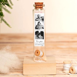Puede incluir: Una botella de vidrio transparente con tapón de corcho, que contiene una bobina de hilo y una tira de tres fotos en blanco y negro. Las fotos son de una pareja. El texto de la tira dice "two years HAPPY COTTON anniversary!". La botella está sobre un pequeño bloque de madera.