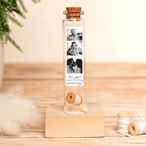 Puede incluir: Un tubo de ensayo de vidrio transparente con tapón de corcho, montado sobre una base cuadrada de madera. En su interior, una tira vertical de tres fotos en blanco y negro sobre un pequeño carrete de madera con hilo blanco y el texto two years HAPPY COTTON anniversary.