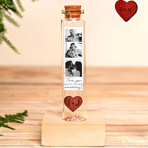 May include: A clear glass bottle with a cork stopper, containing three black and white photos. The bottle sits on a wooden base. A red heart-shaped leather tag with the initials "I+J" is at the bottom. The text reads "Three years HAPPY LEATHER anniversary!"