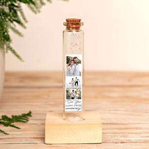May include: A clear glass test tube with a cork stopper, mounted on a light-colored wooden block base. Inside the tube is a vertical strip featuring three photographs of a couple and the text One Year HAPPY PAPER anniversary! in black script.