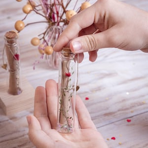 Könnte beinhalten: Eine kleine, klare Glasflasche mit Korken, die eine getrocknete rote Blume und Blätter enthält. Die Flasche wird in den Händen einer Person gehalten, mit einer weiteren Flasche im Hintergrund. Dekorative Herzen und Glitzer sind ebenfalls sichtbar.