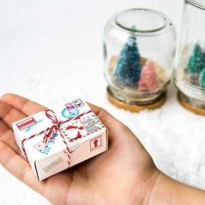 Puede incluir: Una pequeña caja de regalo blanca con hilo rojo y blanco, decorada con sellos y un diseño de Papá Noel. La caja se sostiene en una mano, con dos tarros de vidrio que contienen árboles de Navidad en miniatura en el fondo.