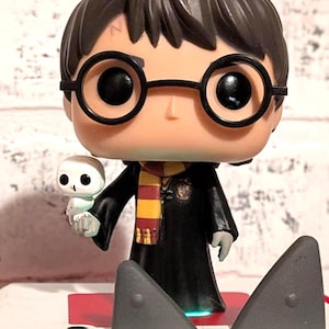 May include: A Funko Pop vinyl figure of Harry Potter, wearing round glasses and a Gryffindor scarf, holding a small white owl. The figure is on a platform with two gray shark fin-shaped elements. The figure is a collectible item.